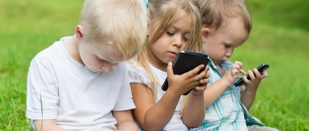 Happy children using smartphones sitting on the grass in the park.